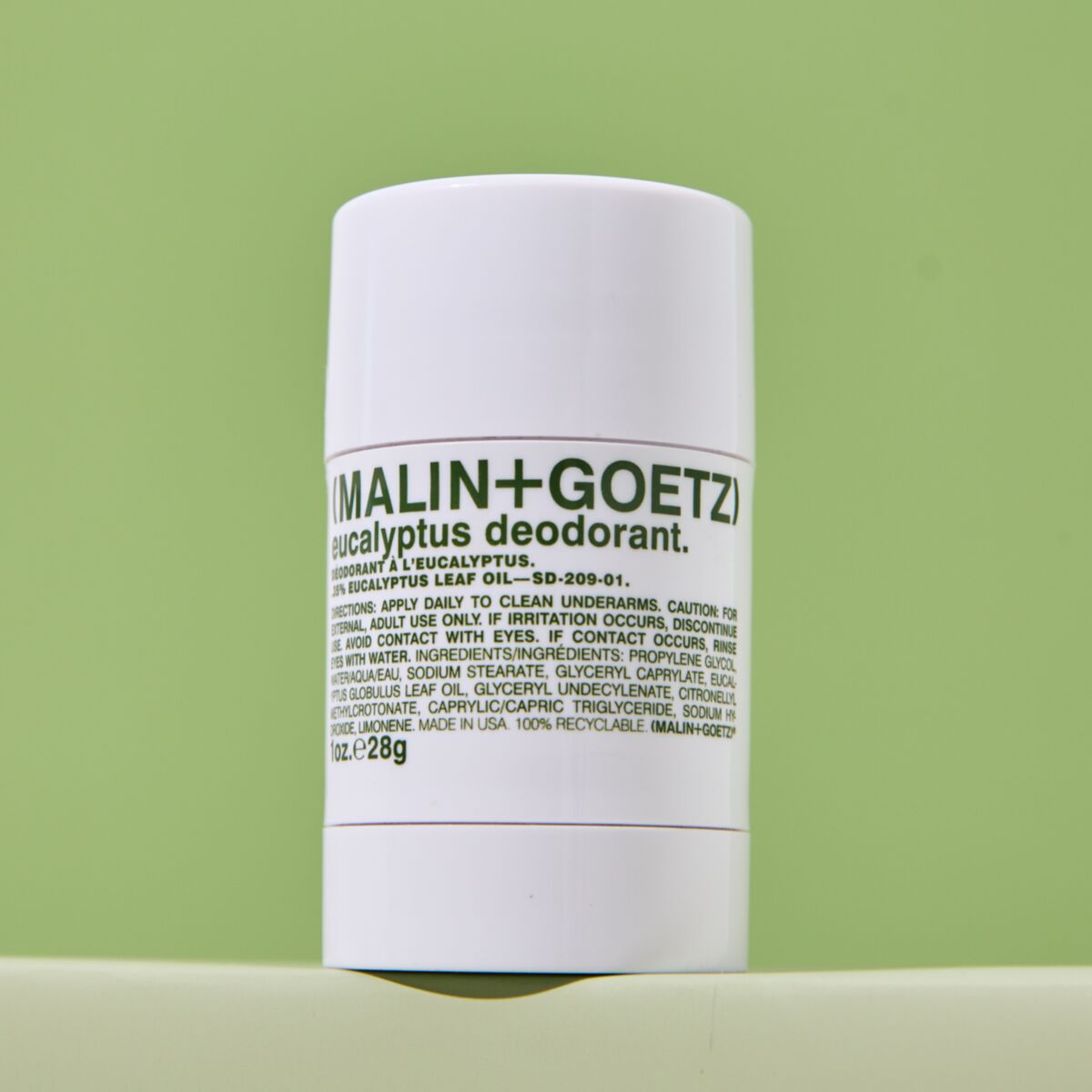 A white cylindrical bottle of Malin+Goetz Eucalyptus Deodorant standing upright against a light green background.