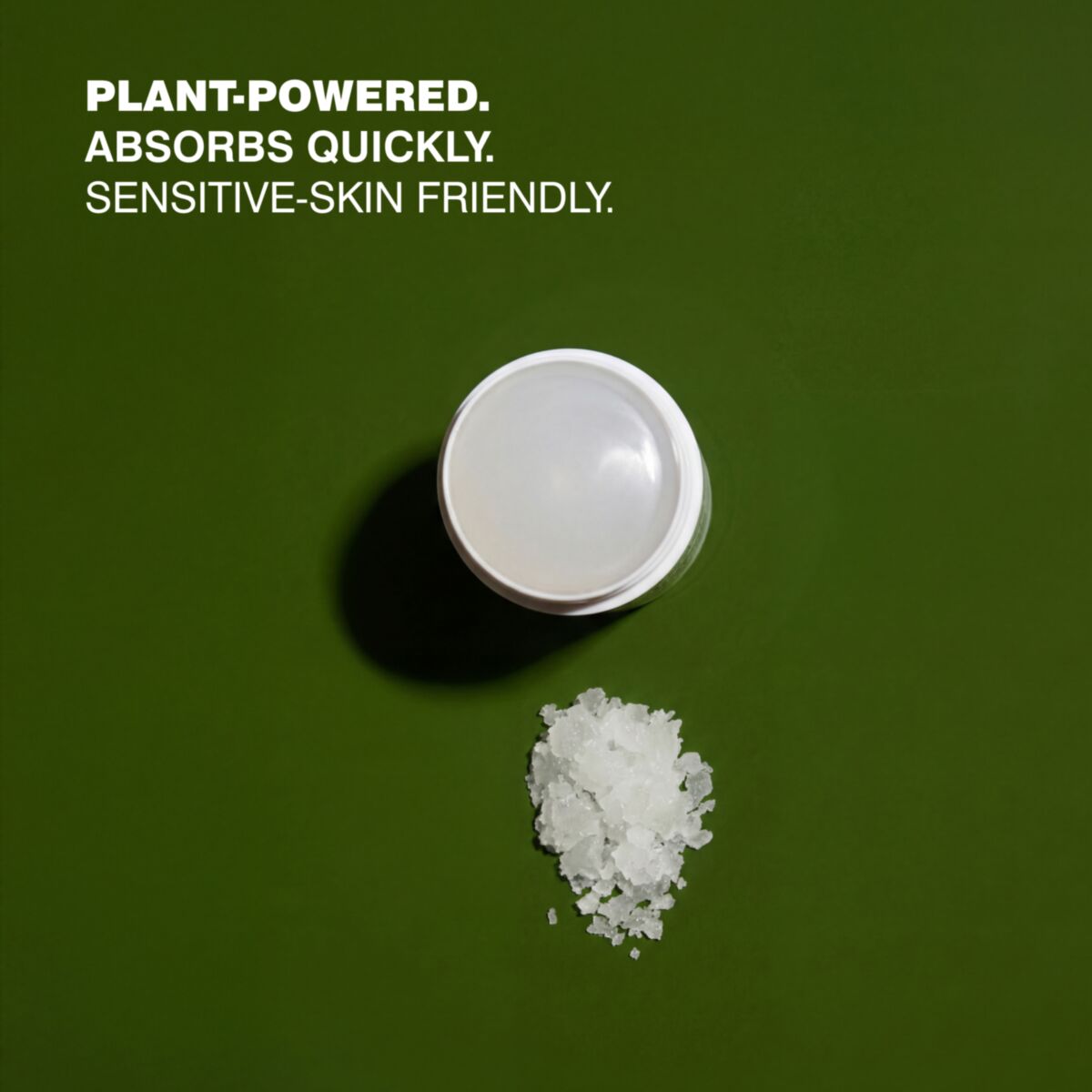 A small white cosmetic jar with its lid off, shown on a dark green background next to a small pile of white granules. White text in the upper left reads: 'PLANT-POWERED.'